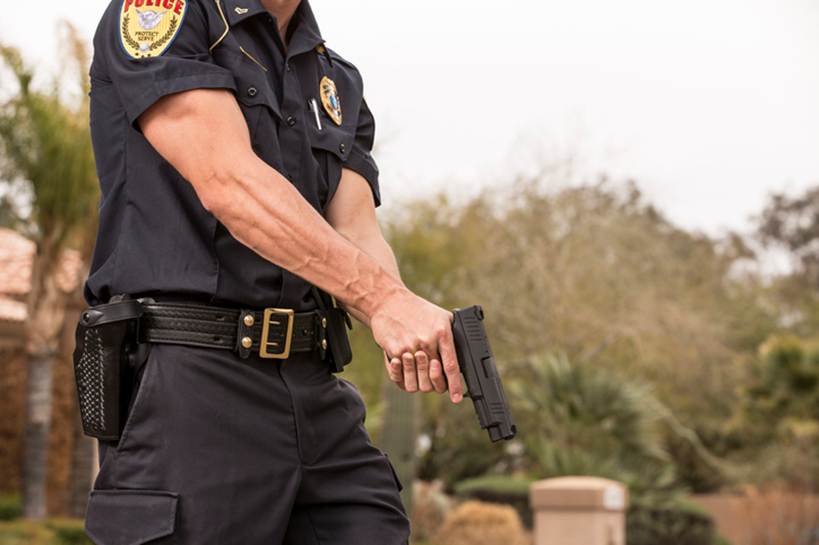 A police officer in uniform stands holding a handgun. The officer's uniform reads "Police, Protect and Serve" on the shoulder patch.